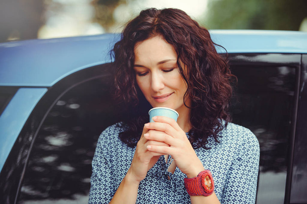 hermosa mujer rizada bebiendo café cerca del coche en un vestido azul
 - Foto, Imagen