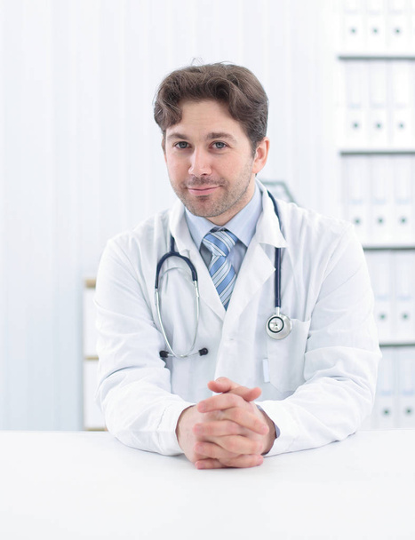 Handsome young doctor in white coat is looking at camera and smiling while standing in office - Photo, image