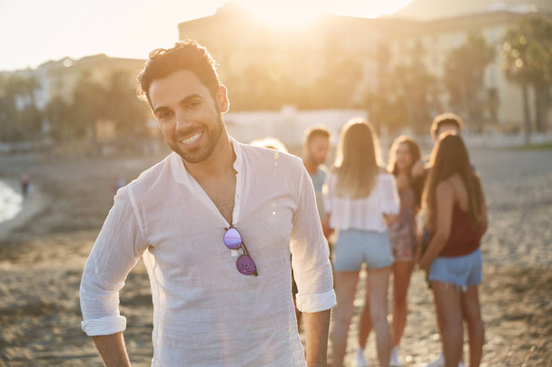 Handsome young man standing on beach with friends smiling - Fotoğraf, Görsel