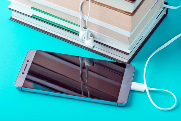 Teléfono con auriculares blancos junto a una pila de libros sobre un fondo azul. Concepto de audiolibros y educación moderna
 - Foto, imagen