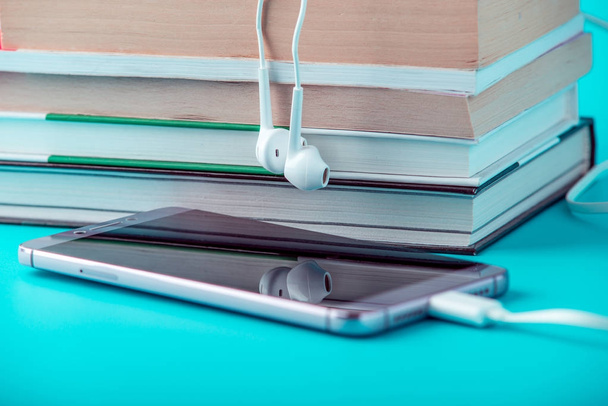Teléfono con auriculares blancos junto a una pila de libros sobre un fondo azul. Concepto de audiolibros y educación moderna
 - Foto, imagen