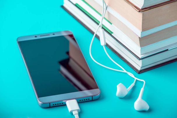 Telefon-val fehér fülhallgató mellett, egy halom könyvet, kék háttérrel. Hangoskönyvek és modern oktatási koncepció - Fotó, kép