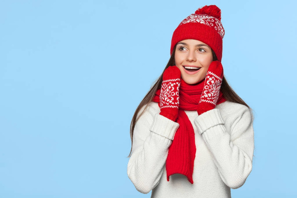 Удивлена, что счастливая женщина в волнении смотрит в сторону. Рождественская девушка в вязаной теплой шляпе и шарфе, изолированные на синем фоне
 - Фото, изображение