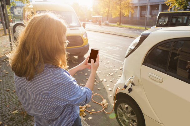 Jeune femme est debout près de la voiture électrique et tenant smartphone
 - Photo, image