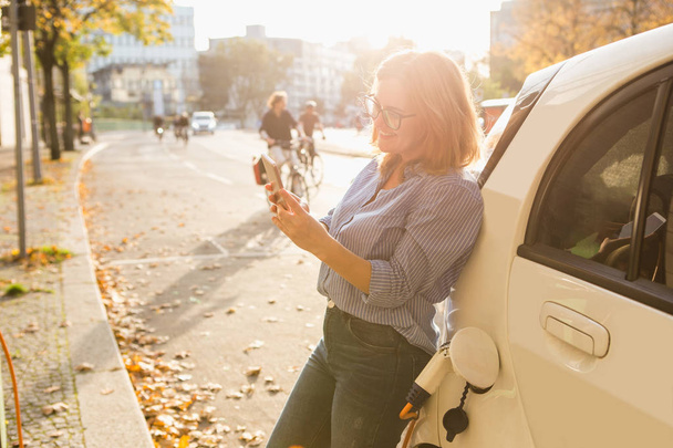 Young woman is standing near the electric car and holding smartphone - Foto, Imagem