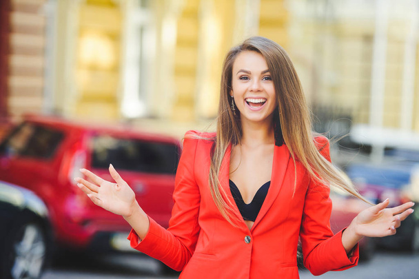 Модная внешность, в жаркий день модель молодой женщины разгуливает по городу в красной куртке, волосах и улыбке на теплом фоне города
 - Фото, изображение