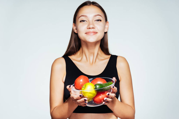 uma jovem atlética em um top preto mantém o controle de sua figura, segura um prato com vegetais úteis e frutas em suas mãos
 - Foto, Imagem