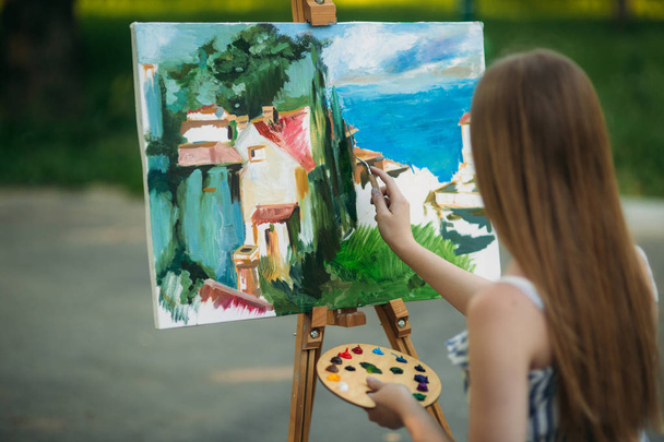 Hermosa chica dibuja un cuadro en el parque usando una paleta con pinturas y una espátula. caballete y lienzo con una imagen
. - Foto, Imagen