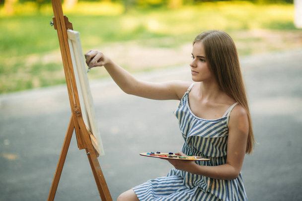 Belle fille dessine une image dans le parc en utilisant une palette avec des peintures et une spatule. Chevalet et toile avec une image
. - Photo, image