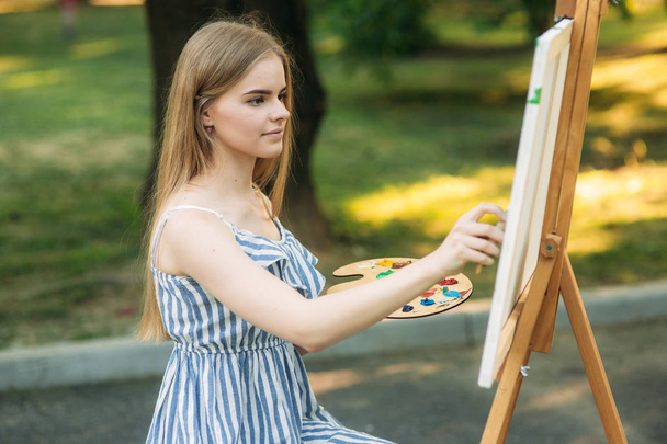 Kaunis tyttö piirtää kuvan puistossa käyttäen paletti maaleilla ja lastalla. Easel ja kankaalle kuva
. - Valokuva, kuva