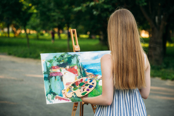 Hermosa chica dibuja un cuadro en el parque usando una paleta con pinturas y una espátula. caballete y lienzo con una imagen
. - Foto, Imagen