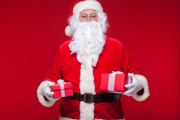 Christmas. Photo Santa Claus giving xmas present and looking at camera, on a red background - Фото, изображение