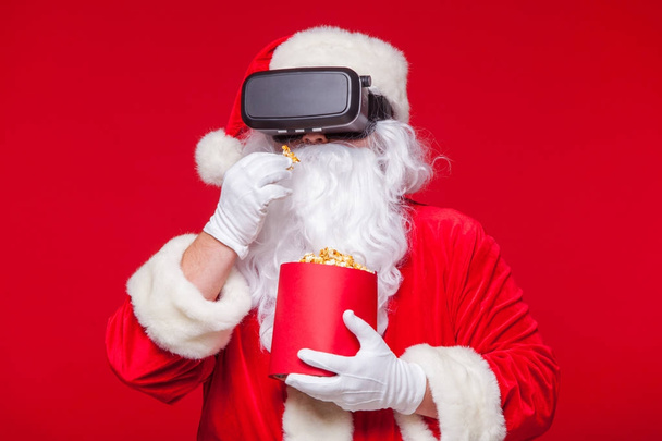 Santa Claus wearing virtual reality goggles and a red bucket with popcorn, on a red background. Christmas - 写真・画像