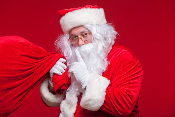 Portrait of Santa Claus with huge red sack keeping forefinger by his mouth and looking at camera - Foto, afbeelding