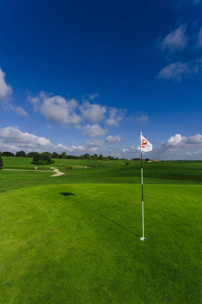Вид на зеленое поле для гольфа, лунку и флаг в яркий солнечный день. Спорт, отдых, отдых и досуг концепция. Летний пейзаж с солнечными лучами
 - Фото, изображение