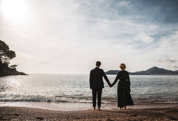 Rear view of happy couple standing against a beach side in summer. Two lovers in vacation in an idyllic nature scene sharing positive feelings and emotions. Magic moments of loving hearts. - Φωτογραφία, εικόνα