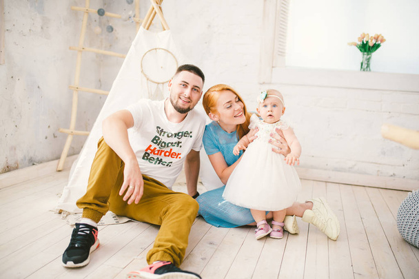 A happy family of three. Mom, dad, child one year old girl in dress play, laugh, smile in bright room. Sunny weather - Photo, Image
