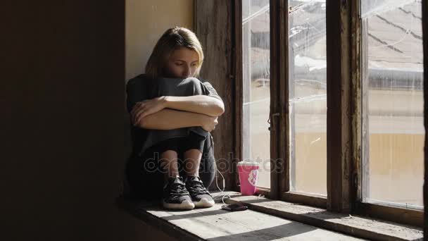 女の子は窓のそばにヘッドフォンで音楽を聞いている間に非常に悲しいように見えます - 映像、動画