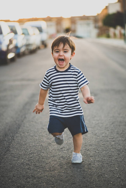 Porträt eines süßen Jungen in kurzen Hosen, der glücklich auf der Straße in Richtung Kamera läuft. - Foto, Bild