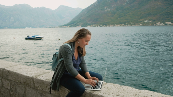 Jonge freelancer vrouw in grijze vest en blauwe rugzak werken op een laptop - Video