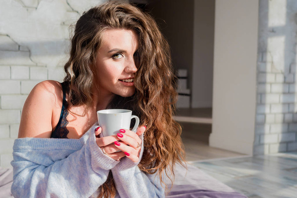 Chica sexy por la mañana con una taza de café en la cama en lencería y suéter. Cabello rizado, mujer rusa sosteniendo una taza con luz natural junto a la ventana en una habitación de matrimonio blanca
. - Foto, Imagen