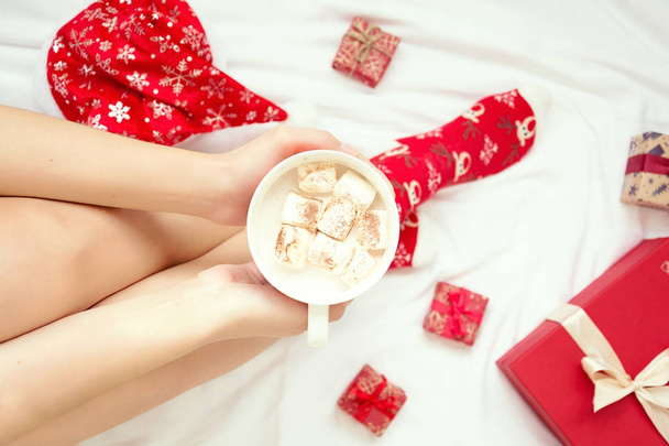 Woman taking mug with hot chocolate and marshmallow on white bed sheet. Overhead shot. - Фото, изображение