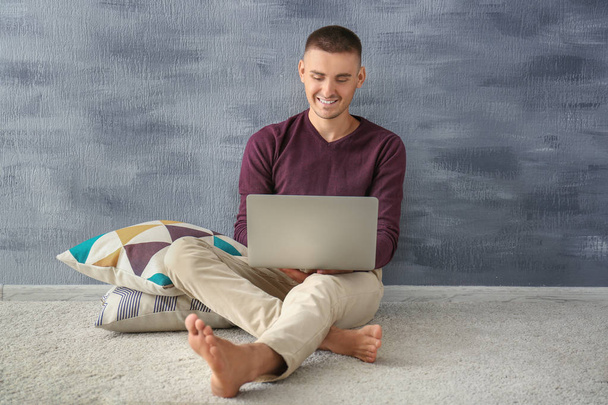 Handsome young man sitting on carpet near grey wall and using laptop - Fotoğraf, Görsel