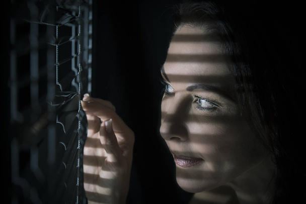 Portrait of a woman standing in darkness looking through blinds  - Foto, Bild