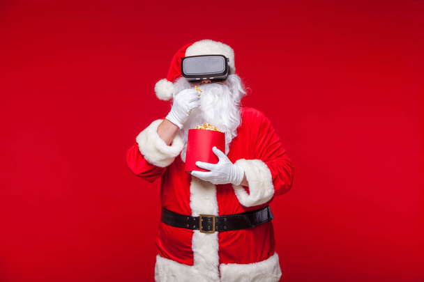 Santa Claus wearing virtual reality goggles and a red bucket with popcorn, on a red background. Christmas - Photo, image