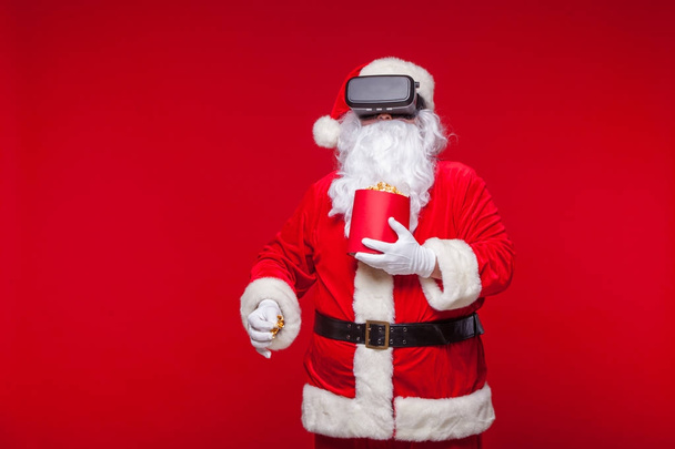 Santa Claus wearing virtual reality goggles and a red bucket with popcorn, on a red background. Christmas - Foto, Imagen