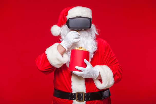Santa Claus visel a virtuális valóság szemüveg és egy piros vödör pattogatott kukorica, piros háttéren. Karácsony - Fotó, kép
