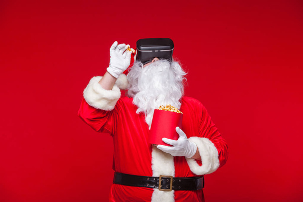 Santa Claus dragen VR-bril en een rode emmer met popcorn, op een rode achtergrond. Kerst - Foto, afbeelding