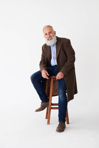 Positive mature bearded man in coat sitting on chair in studio  - Foto, afbeelding