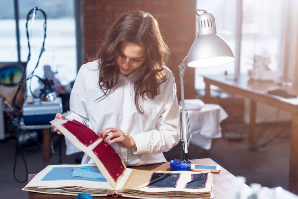 Pretty female tailor choosing a fabric in cloths catalog in studio - Фото, изображение