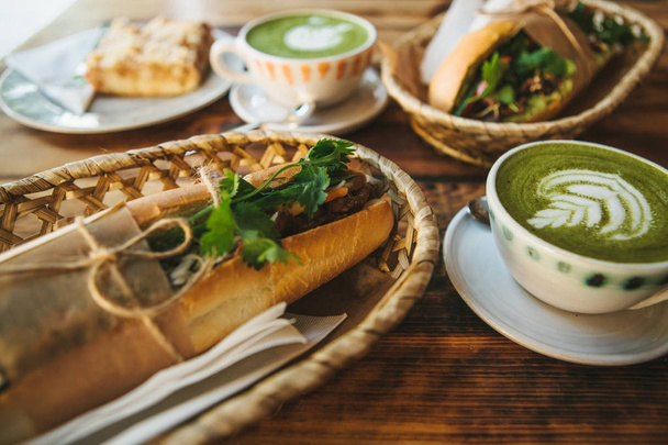 Υγιεινό πρωινό στο εστιατόριο: φλιτζάνι πράσινο τσάι με γάλα, θολή επιδόρπιο και δύο σάντουιτς με λαχανικά και μυρωδικά σε ξύλινο τραπέζι. - Φωτογραφία, εικόνα