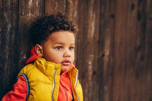 Outdoor portrait of adorable toddler boy posing outside against brown wooden background, wearing orange hoody jacket any bright yellow vest coat - Photo, image