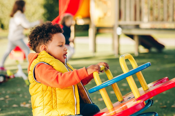 Adorabile bambino di 1-2 anni che si diverte nel parco giochi, bambino con cappuccio arancione e giubbotto giallo
  - Foto, immagini