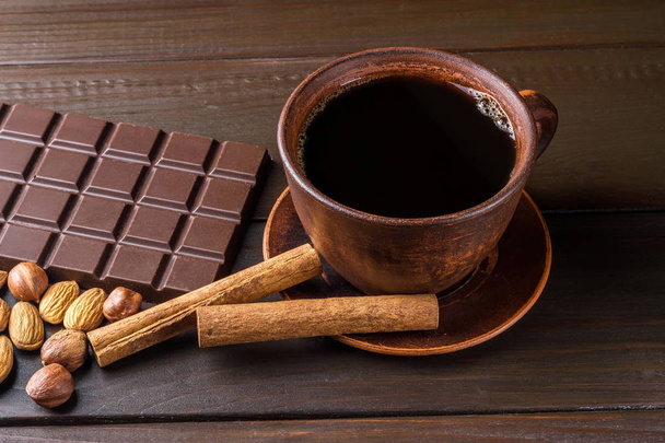 Sıcak siyah kahve kahverengi kil Kupası, koyu çikolata, badem, fındık ve ahşap zemin üzerine tarçın çubuğu içinde. - Fotoğraf, Görsel
