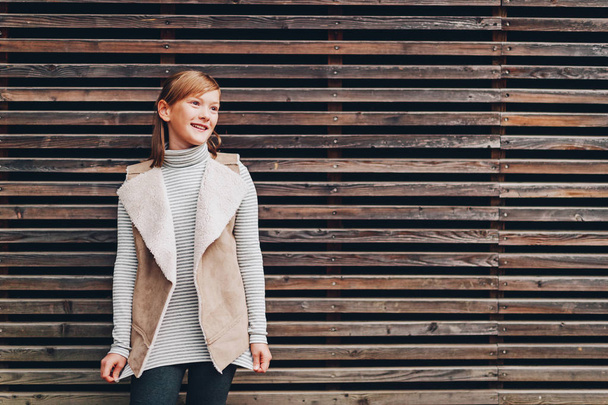 Outdoor-Modeporträt eines hübschen kleinen, 10-jährigen Mädchens, das eine beige Weste trägt und vor braunem Holzhintergrund posiert - Foto, Bild