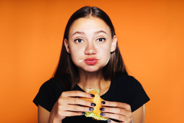 frustrated girl on an orange background eating a burger - Φωτογραφία, εικόνα