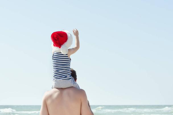 Kind mit Weihnachtsmütze, das auf den Schultern seines Vaters sitzt. Meeresküste. - Foto, Bild