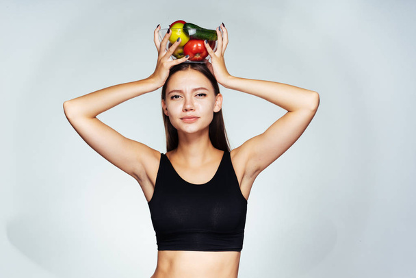 una joven atlética en un top negro hace un seguimiento de su peso, sostiene un plato con verduras útiles y frutas en sus manos
 - Foto, Imagen