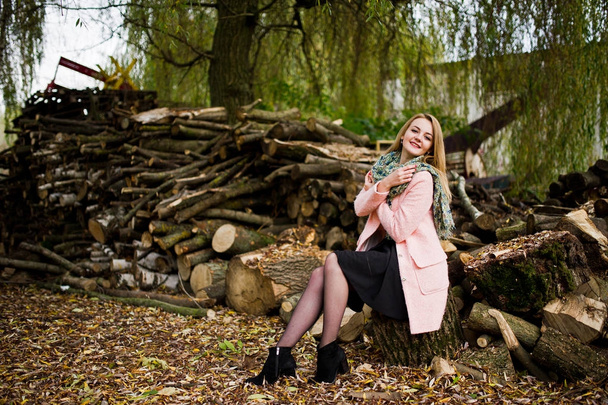 Young blonde girl at pink coat posed against wooden stumps backg - Foto, afbeelding