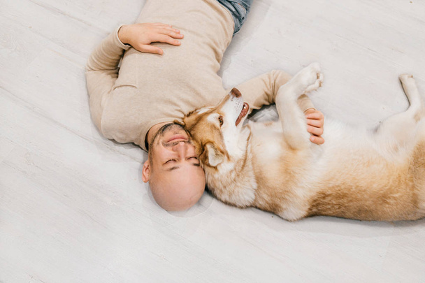 Adult bald man with husky puppy sleeping on floor. Owner with pet together at home. Kind and soulful emotions. Lovely dog resting with young male. Guy with beloved domestic animal hugging each other. - Zdjęcie, obraz