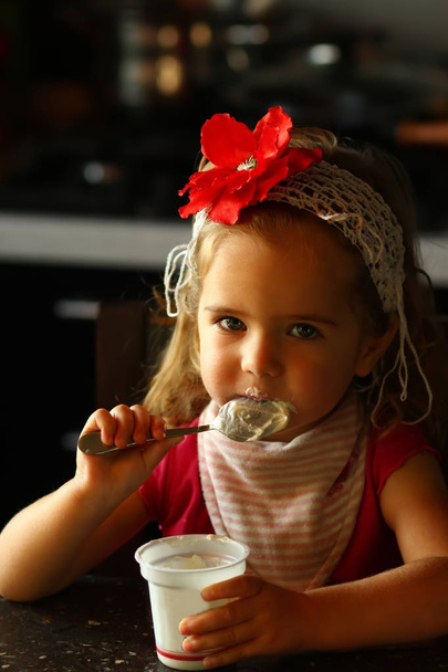 Um ano de idade bebê menina em uma flor vermelha headband comer iogurte por si só para o café da manhã em casa
  - Foto, Imagem
