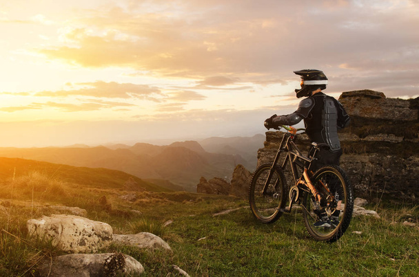 De ruiter in volledige beschermende uitrusting op de mtb fiets rijden richting de zonsondergang in de sunset zonnestralen tegen de achtergrond van de rotsen van de ondergaande zon en wolken - Foto, afbeelding