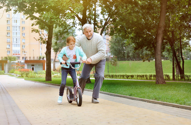 Senior man teaching his granddaughter how to ride kick scooter in park - Foto, Imagem