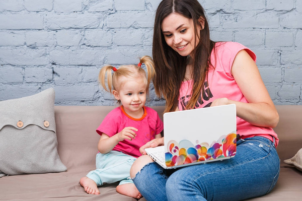 Young mother and daughter of two years old blonde use laptop computer laptop white with bright print sitting on couch indoors - Foto, immagini