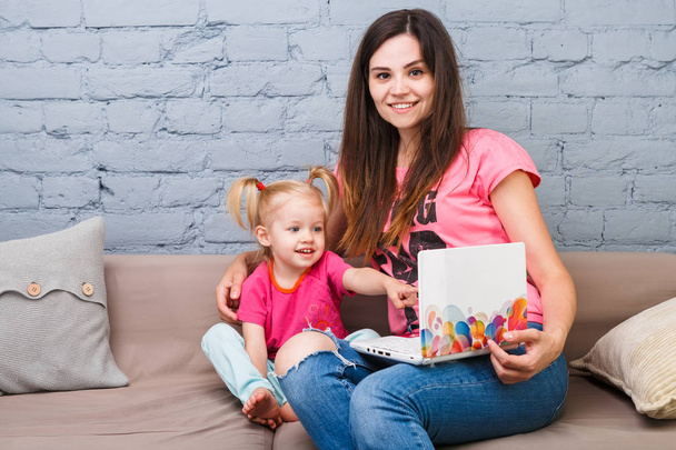 若い母と 2 歳の金髪の娘は室内のソファに座っている明るい印刷でラップトップ コンピューターのラップトップの白を使用します。 - 写真・画像