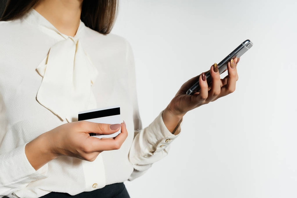 A modern girl in a white shirt is holding a bank card and a smartphone - Photo, Image